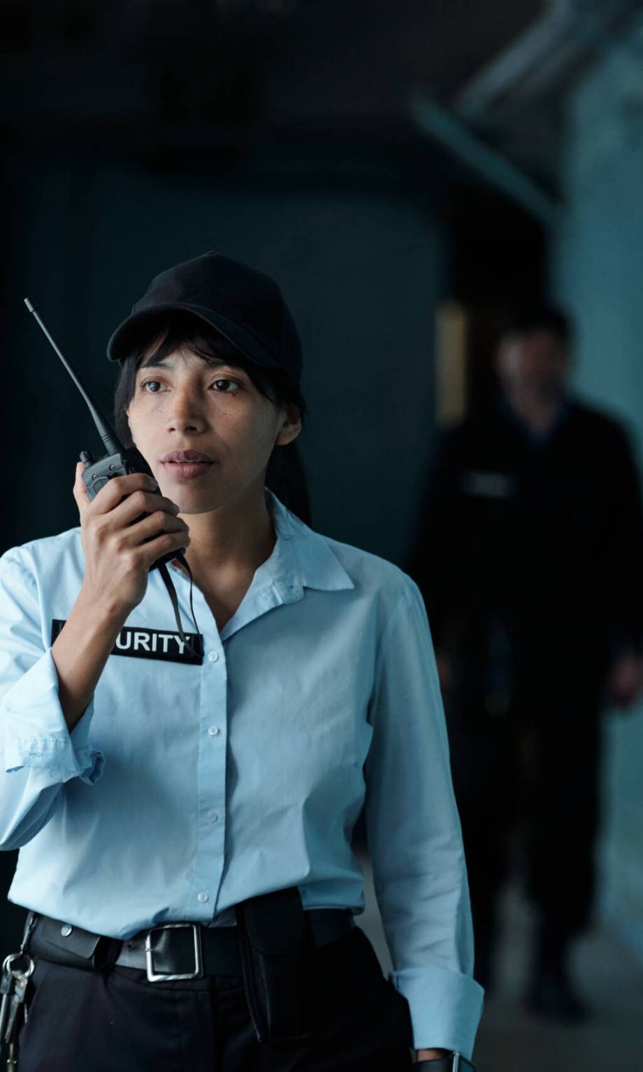 Young female security guard in uniform and cap speaking on walkie-talkie to her colleague while moving along corridor of building